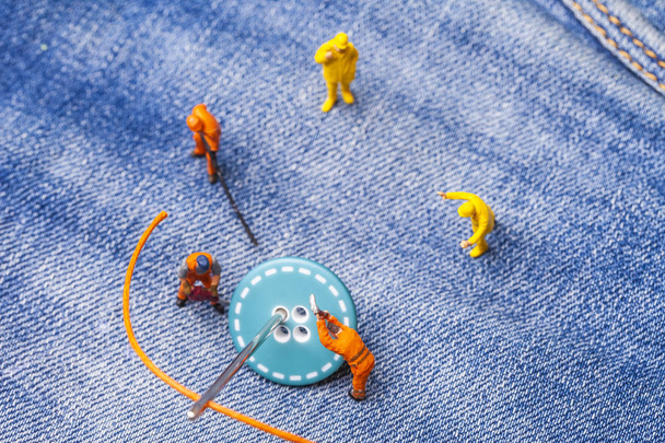 das Konzept für die Modeindustrie. Miniaturarbeiter nähen Knöpfe mit großer Nadel an Jeans. Nahaufnahme. - Foto, Bild