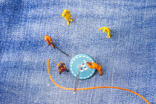 El concepto para la industria de la moda. Los trabajadores en miniatura cosen los botones por la aguja grande a los vaqueros. De cerca.
. - Foto, imagen