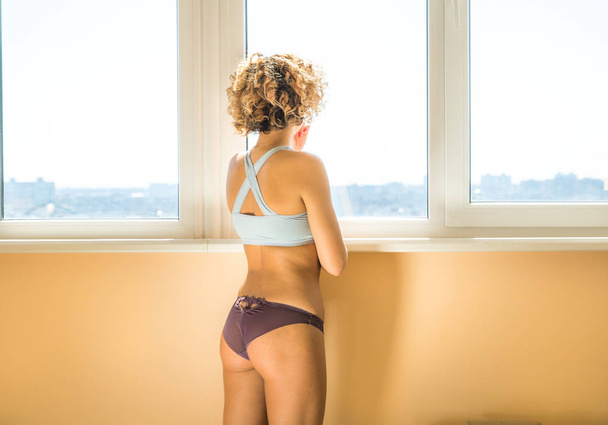 beautiful caucasian curly female in underwear in front of window. orange light background - 写真・画像