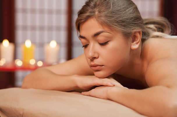 Lovely young woman lying on massage table with her eyes closed, enjoying spa treatment at beauty salon, candles burning on the background. Healthy beautiful female resting at spa on luxury resort - Photo, Image
