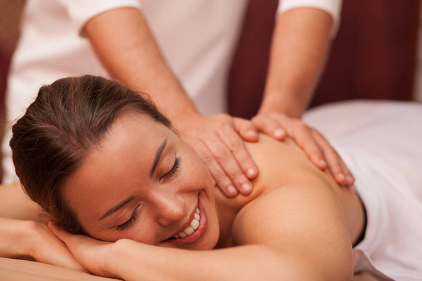 Attractive joyful young woman smiling cheerfully why professional masseur massaging her shoulder. Gorgeous woman relaxing at day spa. Therapist giving soothing massage to female client. Beauty concept - Photo, Image