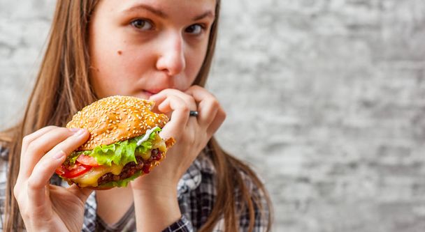 portrait of young teenager brunette girl with long hair eating burger. Girl trying to eat fast food on gray wall background - Foto, immagini