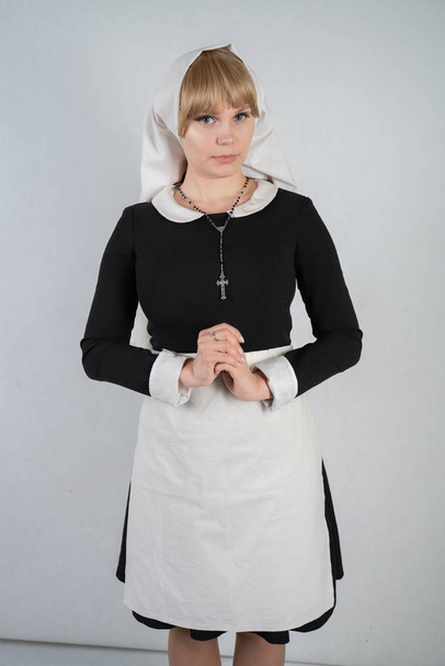 praying caucasian nun stands in a black dress and a white apostolic cap with a cross and an innocent face expression on white studio solid background - Фото, зображення