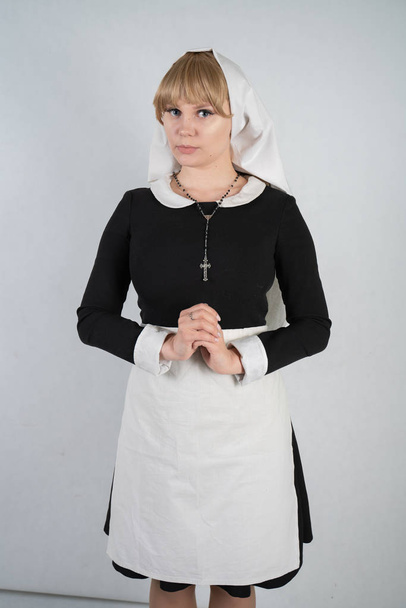 biddende Kaukasische non staat in een zwarte jurk en een witte apostolische dop met een kruis en een onschuldig gezicht expressie op witte studio effen achtergrond - Foto, afbeelding