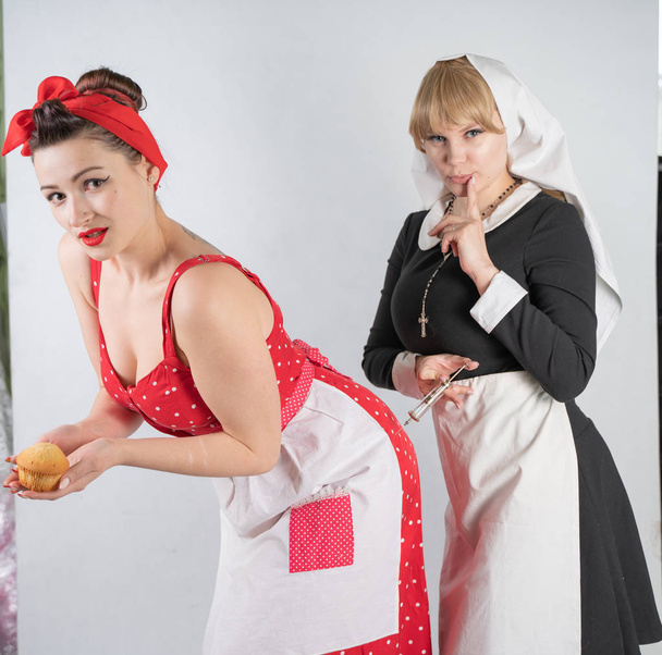 zwei süße Freundinnen verkleidet für eine Kostüm-Cosplay-Party. Ein Mädchen ist als Hausfrau im Pinup-Stil gekleidet, die zweite Frau als Nonne mit Spritze. sie stehen auf weißem Hintergrund. - Foto, Bild