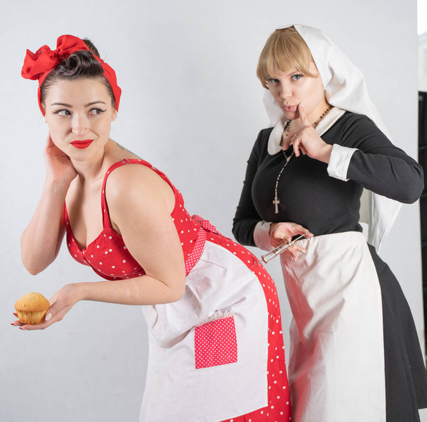 zwei süße Freundinnen verkleidet für eine Kostüm-Cosplay-Party. Ein Mädchen ist als Hausfrau im Pinup-Stil gekleidet, die zweite Frau als Nonne mit Spritze. sie stehen auf weißem Hintergrund. - Foto, Bild