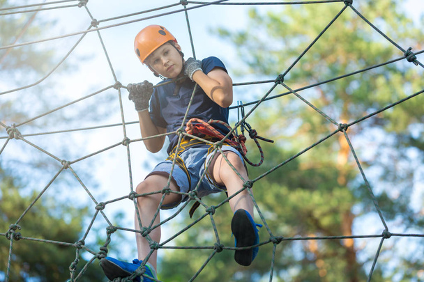 Sporty, young, cute boy in white t shirt spends his time in adventure rope park in helmet and safe equipment in the park in the summer. Active lifestyle concept - Photo, Image