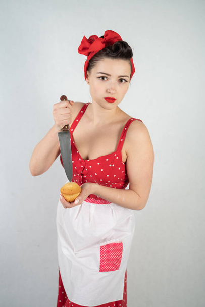небезпечна дівчина-домогосподарка в червоній старовинній сукні з крапкою полоки стоїть з величезним кухонним ножем в руках і сердито радіє на білому твердому студійному фоні
 - Фото, зображення