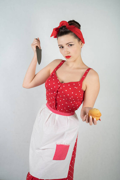 небезпечна дівчина-домогосподарка в червоній старовинній сукні з крапкою полоки стоїть з величезним кухонним ножем в руках і сердито радіє на білому твердому студійному фоні
 - Фото, зображення