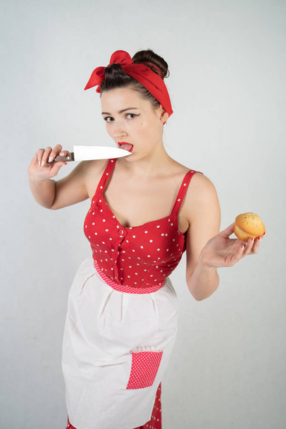 Gefährliche Pin-up-Hausfrau im roten Vintage-Tupfen-Kleid steht mit einem riesigen Küchenmesser in der Hand da und freut sich wütend über weißen soliden Studiohintergrund - Foto, Bild
