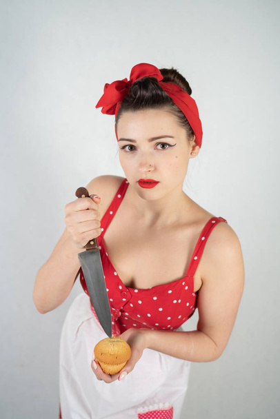 dangerous pin up girl housewife in red vintage polka dot dress stands with a huge kitchen knife in her hands and angrily rejoices on white solid studio background - Fotografie, Obrázek