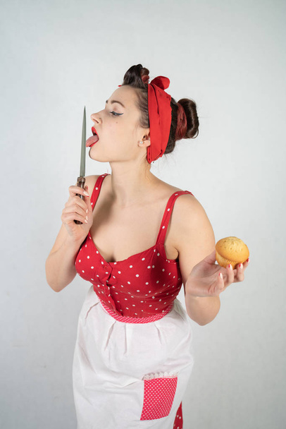 Gefährliche Pin-up-Hausfrau im roten Vintage-Tupfen-Kleid steht mit einem riesigen Küchenmesser in der Hand da und freut sich wütend über weißen soliden Studiohintergrund - Foto, Bild