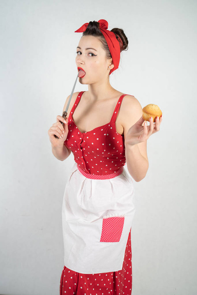 επικίνδυνη pin up κορίτσι νοικοκυρά με φόρεμα κόκκινο πουά ρετρό στέκεται με ένα μαχαίρι τεράστια κουζίνα στα χέρια της και χαίρεται θυμωμένα σε φόντο λευκό στερεό στούντιο - Φωτογραφία, εικόνα