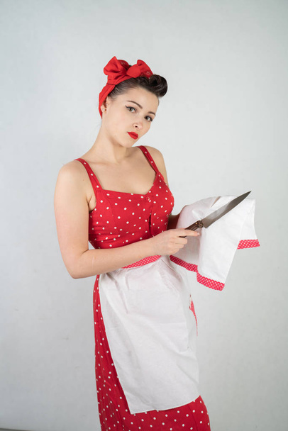 gevaarlijke pin-up girl huisvrouw in rode vintage polka dot jurk staat met een groot keukenmes in haar handen en boos verheugt zich op witte solide studio achtergrond - Foto, afbeelding