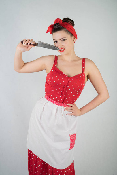 beautiful young girl standing in red polka dot pinup dress and apron, holding a big knife and picking her teeth because the food is stuck, on a white Studio plain background alone - Valokuva, kuva