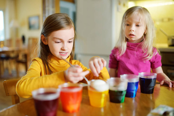 Two cute young sisters dyeing Easter eggs at home. Children painting colorful eggs for Easter hunt. Kids getting ready for Easter celebration. Family traditions. - Photo, image