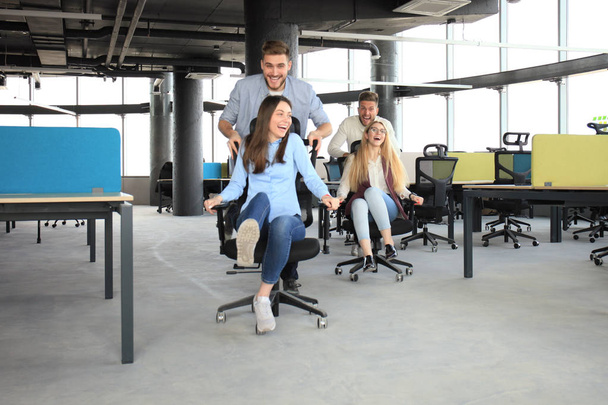 オフィスの椅子の上のレースながら笑みを浮かべて楽しんでスマート カジュアルな服装で若い陽気なビジネス人々. - 写真・画像