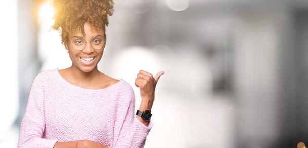 Kaunis nuori afrikkalainen amerikkalainen nainen yllään lasit yli eristetty tausta hymyillen onnellinen kasvot etsivät ja osoittaa sivuun peukalo ylöspäin
. - Valokuva, kuva