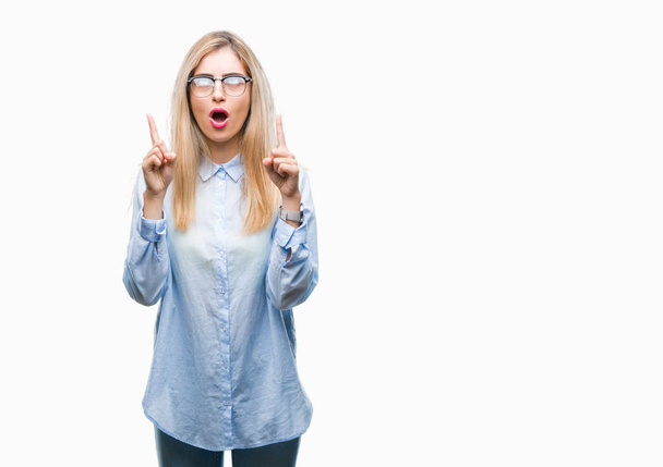Young beautiful blonde business woman wearing glasses over isolated background amazed and surprised looking up and pointing with fingers and raised arms. - Фото, зображення