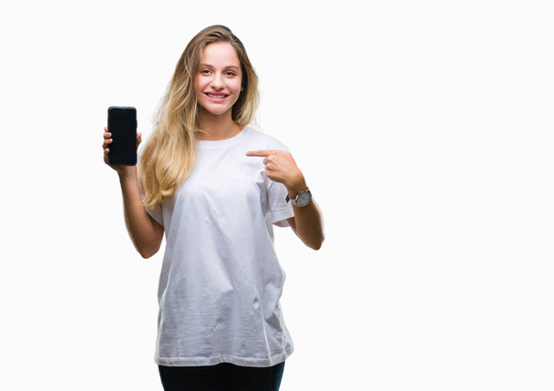Молодая красивая блондинка показывает экран смартфона на изолированном фоне с удивленным лицом, указывающим пальцем на себя
 - Фото, изображение