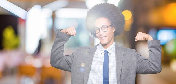 Young african american business man with afro hair wearing glasses showing arms muscles smiling proud. Fitness concept. - Photo, Image