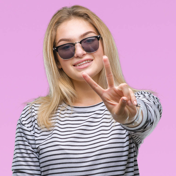 Jeune femme caucasienne portant des lunettes de soleil sur fond isolé souriant avec un sourire heureux clin d'oeil à la caméra faisant signe de victoire. Numéro deux
. - Photo, image