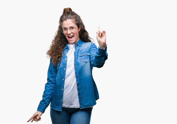 Beautiful young brunette curly hair girl wearing glasses over isolated background pointing finger up with successful idea. Exited and happy. Number one. - Foto, imagen