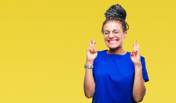 Jeune fille afro-américaine aux cheveux tressés portant des lunettes sur fond isolé souriant croisant les doigts avec espoir et les yeux fermés. La chance et le concept superstitieux
. - Photo, image