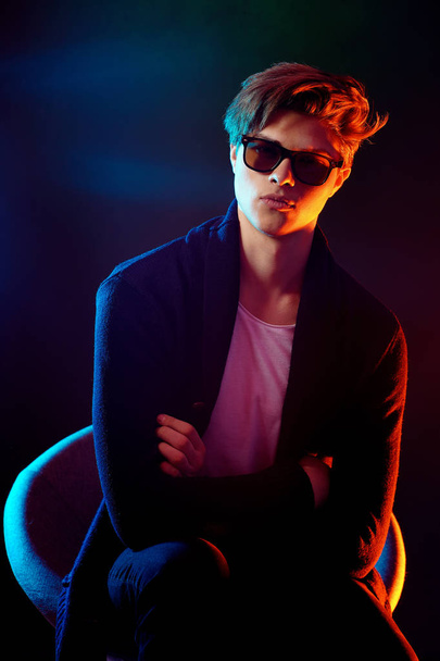Cool stylish man in black jacket and sunglasses. High Fashion male model in colorful bright neon lights posing on black background. Art design concept - Photo, Image