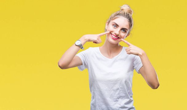 junge schöne blonde Frau trägt weißes T-Shirt über isoliertem Hintergrund, lächelt selbstbewusst und zeigt mit den Fingern Zähne und Mund. Gesundheitskonzept. - Foto, Bild