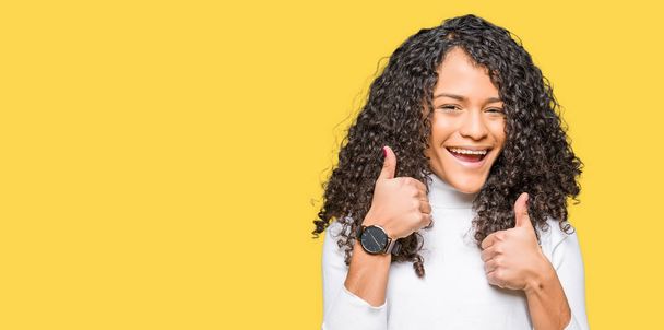 Young beautiful woman with curly hair wearing turtleneck sweater success sign doing positive gesture with hand, thumbs up smiling and happy. Looking at the camera with cheerful expression, winner gesture. - Photo, Image