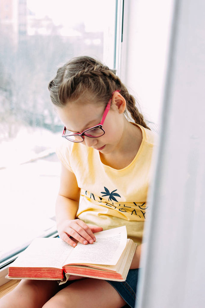Tyttö teini lasit ja letti käsittelyssä kirja ikkunalaudalla hänen huoneessaan
 - Valokuva, kuva