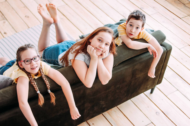 Трое симпатичных улыбающихся детей друзей твитят сидя дома на диване
 - Фото, изображение