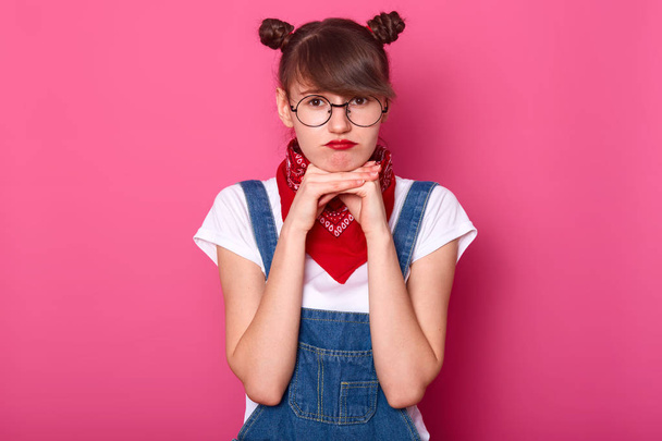 Image of european upset woman with dark hair and bunches, wears overalls, t shirt and bandana. Adorable girl being in bad mood while posing alone in studio, keeps hands under chin. Teenager concept. - Foto, afbeelding