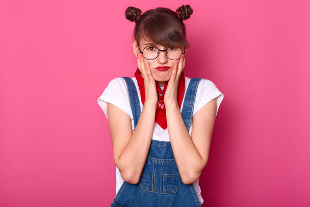 Portrait of offened unhappy teenager girl with pouty lips, keeps hands on cheeks, displeased with everything, feels frustrated and upset, model poses over pink background, copy space for your text. - Foto, afbeelding