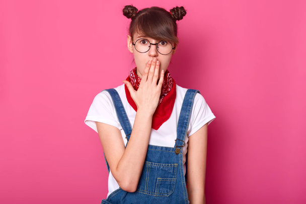 Image of surprised dark haired teenager with funny banches, covers mouth with hand, wears t shirt, overalls, glasses and bandana, looks with wide open eyes, hears shocking news, isolated on pink wall. - Foto, afbeelding