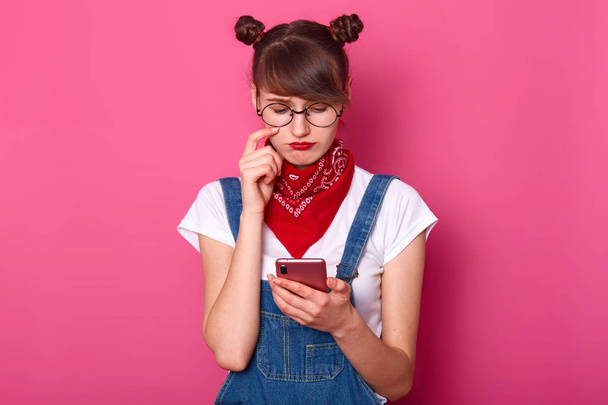 Young adorable woman wears denim overalls, casual white t shirt, red bandana on neck and rounded spectacles, with bunches feels sad while reading sms on her mobile phone, isolated over pink background - Фото, изображение