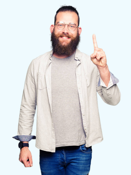 Young blond man wearing glasses showing and pointing up with finger number one while smiling confident and happy. - Foto, immagini