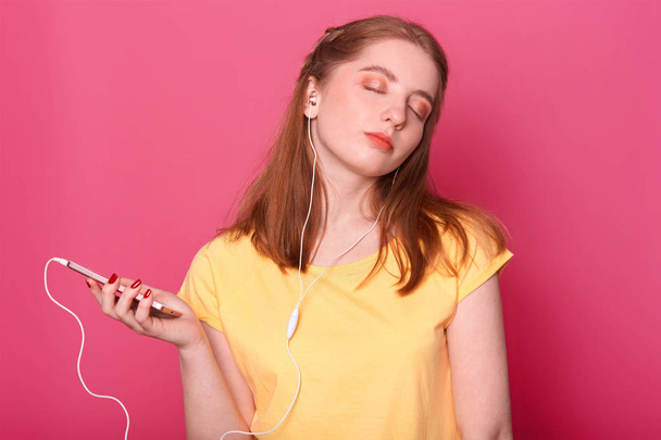 Dreamy female with pensive expression and closeed eyes, has modern headphones, listen to music, spends leisure time alone, poses against pink background with blank copy space for your avertisment. - Фото, изображение