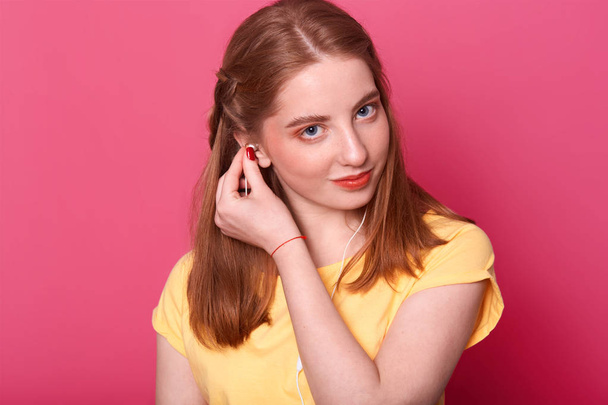 Attractive young girl puts earphones, ready to listen to music, looks derictly at camera, wears bright yellow t shirt, has brown straight hair, spend free time alone. People and entairtaiment concept. - Фото, зображення
