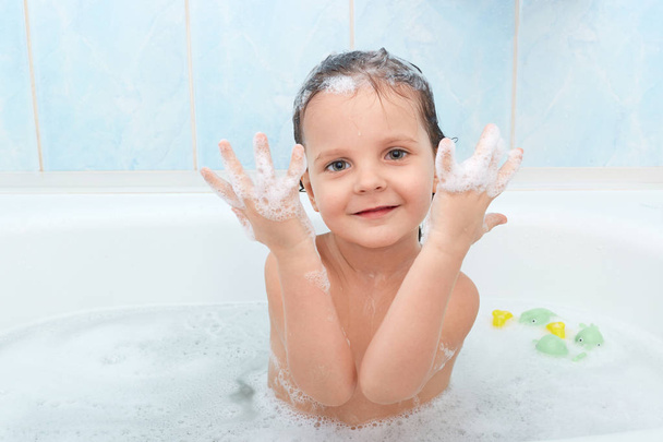 Small adorable child has funny expression, taking bath with plesure, plaing in hot water and shows hands with foam, poses in bathroom against blue wall. Playful little kid glad to be photographed. - Fotoğraf, Görsel