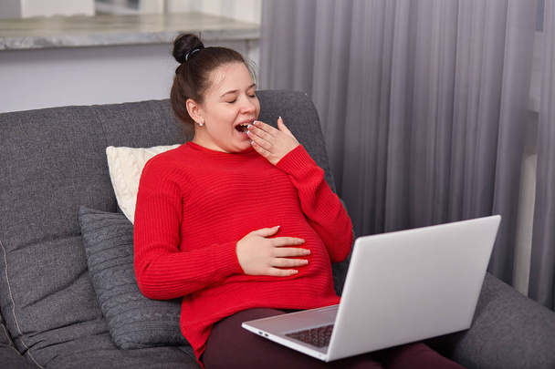 Attractive pregnant female sits on sofa, keeps hand on belly, anticipates for baby, yawns as tired of watching videos in Internet, dressed in sweater, with bunch. Pregnancy, technology concept - Fotó, kép