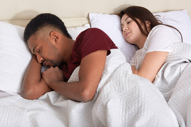 Крупный план пары, спящей в постели, отдыхающей после тяжелого дня, одетой в футболки, лежащей с закрытыми глазами, пытающейся заснуть, муж обиженный женой и повернутый спиной. Концепция отношений и людей
 - Фото, изображение