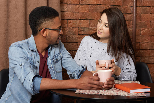 Romantic relaxed couple sit at table in cafe, look at each other, hold cups of coffee or tea in hands, being of different race, enjoy togetherness, spend weekend together. People, relationship concept - Fotoğraf, Görsel