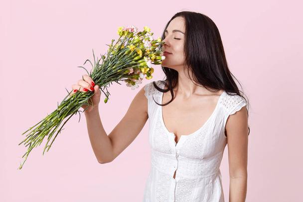 Portrait of gentle dark haired young lady, dressed in white dress, happy to recieve gift on birthday, stands with closed eyes, smels bouquet of beautiful flowers, models poses over rosy background. - Photo, image
