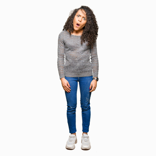 Young beautiful woman with curly hair wearing grey sweater In shock face, looking skeptical and sarcastic, surprised with open mouth - Photo, Image