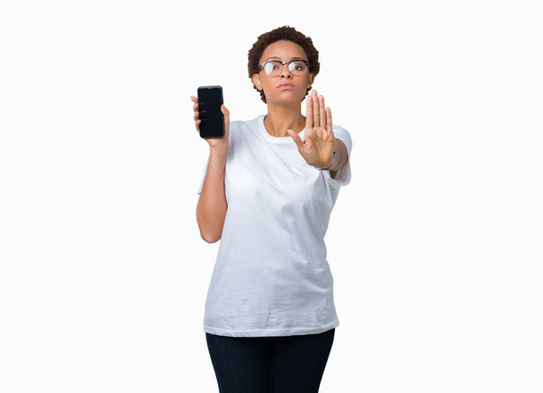 Mujer afroamericana joven mostrando la pantalla del teléfono inteligente sobre un fondo aislado con la mano abierta haciendo señal de stop con expresión seria y segura, gesto de defensa
 - Foto, imagen