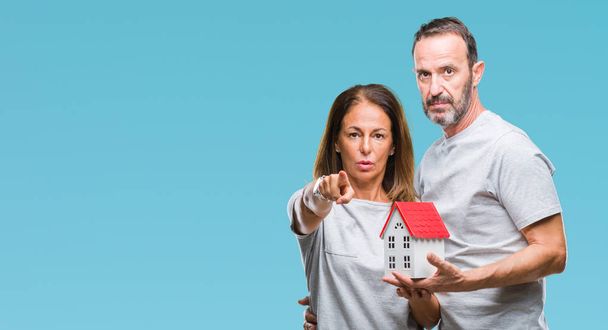 Μέση ηλικία Ισπανόφωνος casual ζευγάρι αγοράζοντας νέο σπίτι πέρα από το απομονωμένο υπόβαθρο, δείχνοντας με το δάχτυλο στη φωτογραφική μηχανή και σε εσάς, το χέρι συνδεθείτε, θετική και σίγουρη κίνηση από το μέτωπο - Φωτογραφία, εικόνα