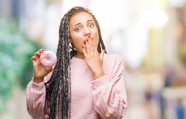 Young african american girl eating pink donut over isolated background cover mouth with hand shocked with shame for mistake, expression of fear, scared in silence, secret concept - Photo, Image