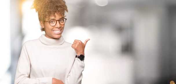 Beautiful young african american woman wearing glasses over isolated background smiling with happy face looking and pointing to the side with thumb up. - Foto, Imagem